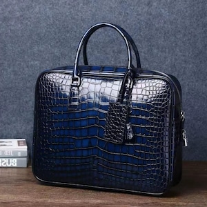 May include: A navy blue briefcase with a crocodile skin texture. The bag has a top handle, a small tag, and a zipper closure. The briefcase is sitting on a wooden surface.