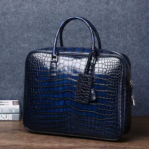 May include: A blue leather briefcase with a crocodile skin texture. The bag has a top handle, a zip closure, and a small tag. The briefcase is sitting on a wooden surface.