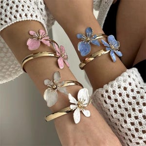 May include: Gold-tone metal bracelets with floral accents in pink, blue, and white. Each bracelet features a delicate flower design with a gold center. The bracelets are worn on the arm and are a fashion accessory.