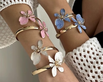 May include: Gold-tone metal bracelets with floral accents in pink, blue, and white. Each bracelet features a delicate flower design with a gold center. The bracelets are worn on the arm and are a fashion accessory.