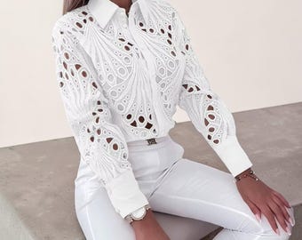 May include: White button-down shirt with intricate cut-out detailing on the sleeves and bodice. The shirt has a classic collar and cuffs. The model is wearing white pants and a watch.