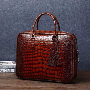May include: A brown leather briefcase with a crocodile skin texture. The bag has a top handle, a zipper closure, and a small tag. The briefcase is sitting on a wooden surface.