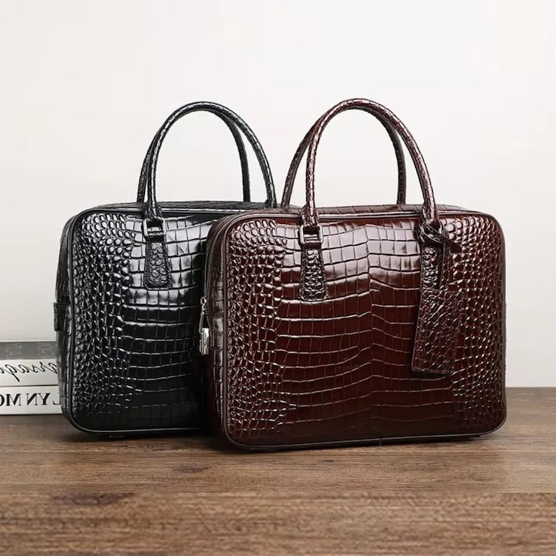 May include: Two briefcases with a crocodile skin texture. One is black, the other is brown. Both have top handles and a rectangular shape. The briefcases are sitting on a wooden surface.