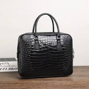 May include: A black briefcase with a glossy, crocodile-skin texture. The bag has two short handles and a zipper closure. The briefcase is rectangular and sits on a wooden surface.