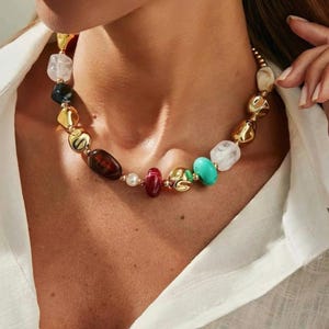Peut inclure: Un collier de perles colorées avec une variété de formes et de couleurs. Le collier comprend des accents métalliques dorés et des perles dans les tons de blanc, noir, marron, rouge, vert et turquoise. Le collier est porté sur une chemise blanche.