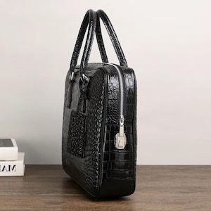 May include: Black briefcase with a crocodile skin texture. The bag has two handles, a silver zipper, and a small pocket on the front. The briefcase is sitting on a wooden surface.