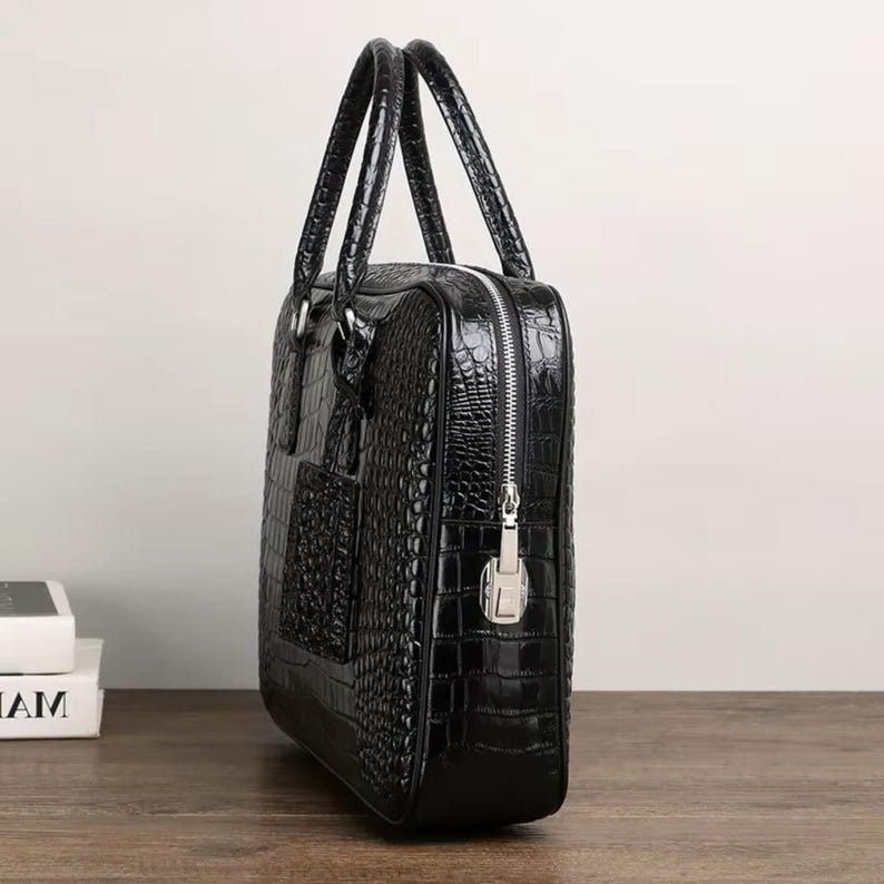 May include: Black briefcase with a crocodile skin texture. The bag has two handles, a silver zipper, and a small pocket on the front. The briefcase is sitting on a wooden surface.