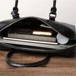 May include: Black leather briefcase with a textured crocodile pattern. The open bag reveals a laptop, notebook, wallet, and other items. The briefcase has a zipper closure and a handle.