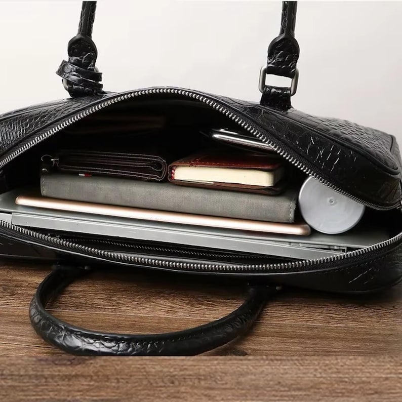 May include: Black leather briefcase with a textured crocodile pattern. The open bag reveals a laptop, notebook, wallet, and other items. The briefcase has a zipper closure and a handle.
