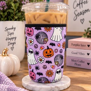May include: A reusable iced coffee cup sleeve featuring a light purple background with a repeating pattern of orange pumpkins, white ghosts wearing sunglasses, disco balls, black bats, spiders, orange flowers, and candy corn, finished with black trim.