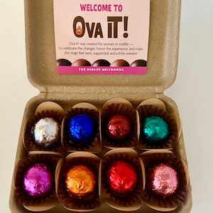 May include: An open cardboard container holding eight foil-wrapped chocolates in various colors. A pink card inside reads "WELCOME TO Ova It!" and describes the product's purpose. The chocolates are in brown paper cups.