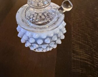May include: A clear glass jar with a lid and a handle. The jar has a textured, hobnail-like design on the bottom and a decorative lid. The handle is a small, round knob. The jar is sitting on a dark brown surface.