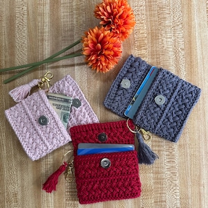 May include: Three rectangular crochet pouches in light pink, dark red, and slate gray. Each pouch features a textured cable-knit pattern, a silver magnetic snap closure, a gold-tone lobster clasp, and a matching yarn tassel attached to the side.