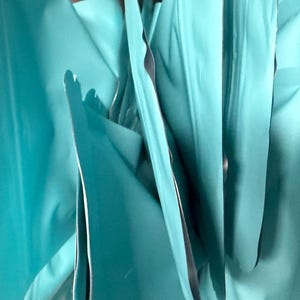 May include: A collection of multiple matte turquoise stand-up pouches arranged in a vertical, overlapping formation. Each pouch features a smooth, opaque surface with sealed edges, creating a series of folds and shadows in a bright teal hue.