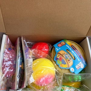 K&ouml;nnte beinhalten: Ein Pappkarton mit verschiedenen &Uuml;berraschungsspielzeugen in durchsichtiger Plastikverpackung. Enthalten sind eine leuchtend gelb-rosa Kugel, eine rote Kugel und ein runder beiger Beh&auml;lter mit einem blauen Etikett mit der Aufschrift Rainbow Mystery.