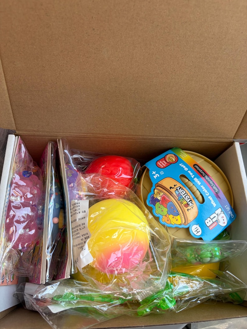 K&ouml;nnte beinhalten: Ein Pappkarton mit verschiedenen &Uuml;berraschungsspielzeugen in durchsichtiger Plastikverpackung. Enthalten sind eine leuchtend gelb-rosa Kugel, eine rote Kugel und ein runder beiger Beh&auml;lter mit einem blauen Etikett mit der Aufschrift Rainbow Mystery.