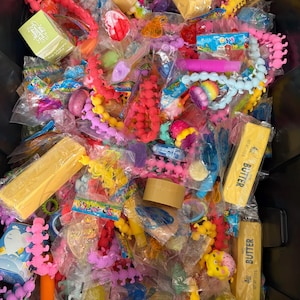 K&ouml;nnte beinhalten: Eine Sammlung verschiedener kleiner Spielzeuge und Scherzartikel in individuellen Kunststoffverpackungen, darunter leuchtend rosa und gelbe raupenf&ouml;rmige Fidgets, violette Pop-Tubes und rechteckige gelbe Schachteln mit der Aufschrift 4oz butter.