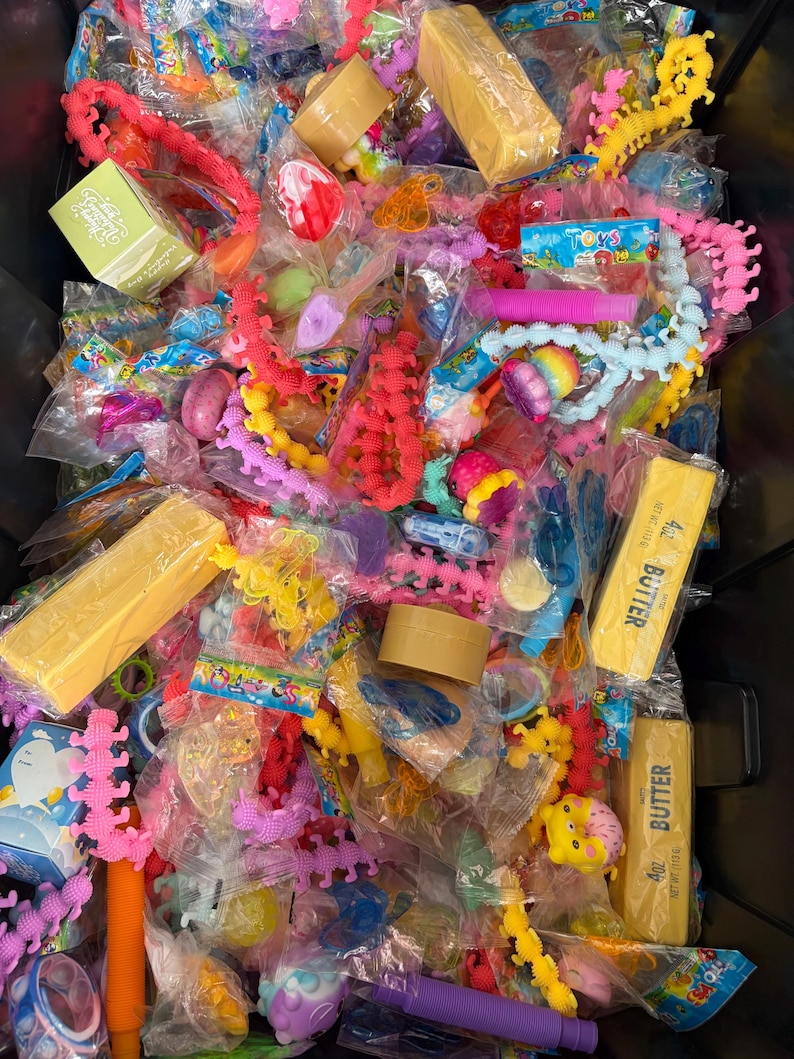 K&ouml;nnte beinhalten: Eine Sammlung verschiedener kleiner Spielzeuge und Scherzartikel in individuellen Kunststoffverpackungen, darunter leuchtend rosa und gelbe raupenf&ouml;rmige Fidgets, violette Pop-Tubes und rechteckige gelbe Schachteln mit der Aufschrift 4oz butter.