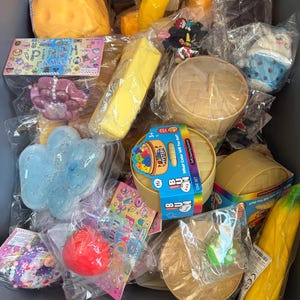 May include: A collection of colorful, packaged squishy toys, including a cheese wedge, banana, and cloud-shaped items. Other items include a round container, a box of crayons, and a pink box with the text "PINCH FAMILY".