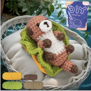 May include: A handmade crochet otter toy in a white basket. The otter is brown, white, and tan, lying on a green and yellow crocheted blanket. A DIY crochet kit is visible, along with several yarn skeins in yellow, brown, green, and tan.