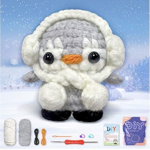 May include: A handmade crochet penguin wearing a white earmuff and scarf. The penguin has black eyes, an orange beak, and gray and white body. The image includes yarn, crochet hooks, and a DIY crochet kit, suggesting a craft project.