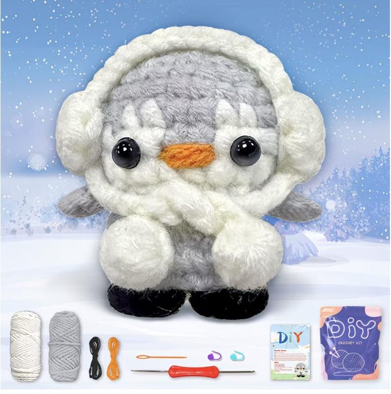 May include: A handmade crochet penguin wearing a white earmuff and scarf. The penguin has black eyes, an orange beak, and gray and white body. The image includes yarn, crochet hooks, and a DIY crochet kit, suggesting a craft project.