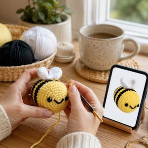 Peut inclure: Un crochet en forme d'abeille jaune et noire avec des ailes blanches est en cours de r&eacute;alisation avec un crochet. Un smartphone affiche un motif d'abeille similaire. Des pelotes de laine jaunes, noires et blanches sont dans un panier.