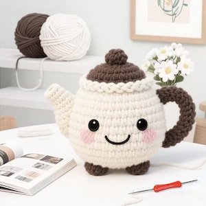 May include: A handmade crochet teapot with a smiling face, brown handle, and lid. The teapot is white with brown accents and pink cheeks. Two balls of yarn, one brown and one white, are in the background. A small red and silver crochet hook is in the foreground.