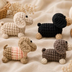 May include: Four crocheted dachshund dog figurines in various colours: beige, black, brown, and a brown and pink striped one. Each dog has small black eyes and tan-coloured paws. The dogs are handmade and sit on a neutral-coloured surface.
