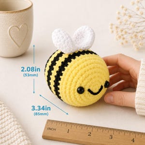 Peut inclure: Une abeille faite &agrave; la main, crochet&eacute;e, avec des rayures jaunes et noires, des ailes blanches et un visage souriant. L'abeille mesure 8,5 cm de long et 5,3 cm de haut. Une r&egrave;gle en bois est au premier plan.