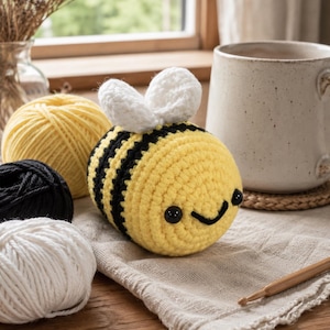 May include: A handmade crochet bee with yellow and black stripes, white wings, and a smiling face. The bee is placed on a linen cloth, with balls of yellow, black, and white yarn in the background. A mug and a crochet hook are also visible.