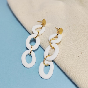 Puede incluir: Pendientes colgantes ovalados blancos con detalles dorados. Los pendientes presentan tres óvalos blancos conectados, suspendidos de un perno dorado. Los pendientes se muestran sobre un fondo azul claro y beige.