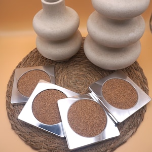 May include: Four square metal coasters with cork centers are arranged on a woven mat, alongside two decorative, stacked, beige ceramic vases. The coasters have a modern, minimalist design, suitable for home decor.