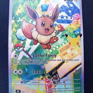 May include: A holographic Eevee trading card featuring the character in the centre with 60 HP. The card displays the text Call for Family and Tackle, along with a 25th-anniversary logo. The background is a colourful, abstract geometric design.