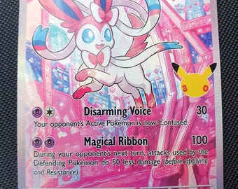 Puede incluir: Una carta coleccionable de Sylveon con fondo rosa que muestra al personaje de ojos azules con antenas en forma de cinta. La carta indica 110 HP, los ataques Voz Cautivadora y Cinta Mágica, y un pequeño logotipo amarillo de Pikachu.