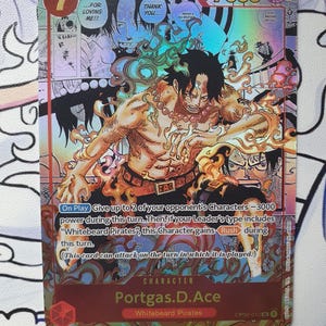 May include: A holographic trading card featuring the character Portgas D. Ace from the "One Piece" series. The card has a vibrant design with a central illustration of Ace surrounded by flames, with text that includes "7000" and "Whitebeard Pirates".