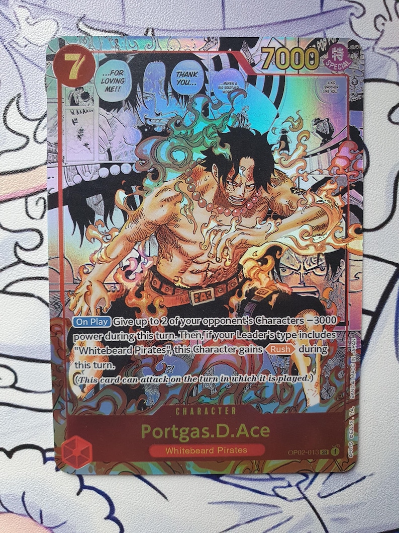 May include: A holographic trading card featuring the character Portgas D. Ace from the "One Piece" series. The card has a vibrant design with a central illustration of Ace surrounded by flames, with text that includes "7000" and "Whitebeard Pirates".