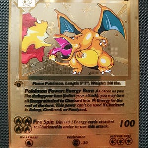 May include: A gold-coloured Charizard trading card featuring the character breathing fire. The card displays 120 HP, the Flame Pokémon description, Energy Burn power, Fire Spin attack with 100 damage, and various game statistics in black text.