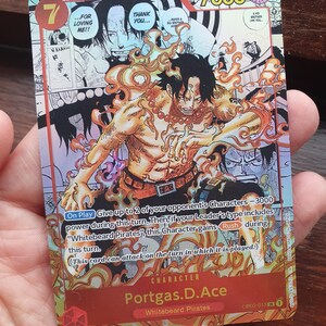 May include: A collectible trading card featuring the character Portgas D. Ace from the "One Piece" series. The card has a vibrant design with a fiery aura around the character, with the number 7 and the text "7000" visible. The card is held in a hand.