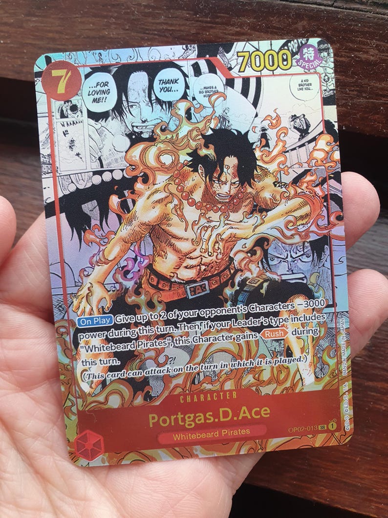May include: A collectible trading card featuring the character Portgas D. Ace from the "One Piece" series. The card has a vibrant design with a fiery aura around the character, with the number 7 and the text "7000" visible. The card is held in a hand.