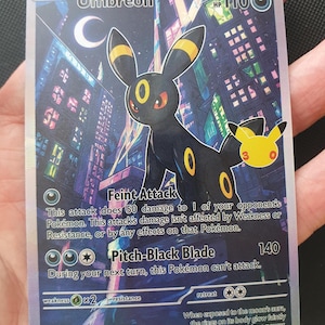 May include: A holographic Umbreon trading card featuring a black creature with yellow rings against a dark city skyline at night. The card displays 110 HP, Feint Attack, and Pitch-Black Blade moves, with a 25th-anniversary logo and 2026 copyright text.