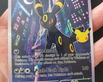 Puede incluir: Una carta coleccionable holográfica de Umbreon que muestra una criatura negra con anillos amarillos sobre un paisaje urbano nocturno. La carta indica 110 HP, los ataques Feint Attack y Pitch-Black Blade, un logo de aniversario y el texto 2026.