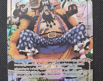Puede incluir: Una carta coleccionable holográfica de One Piece con Marshall D. Teach. La carta muestra un coste de 10, un poder de 12000 y texto que detalla las habilidades Blocker y Activate:Main. El personaje aparece en una pose sentada con un sombrero pirata.