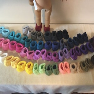May include: A collection of colorful doll-sized knitted booties in various colors, including white, pink, yellow, green, blue, purple, and gray. The booties are arranged in a semi-circle on a white surface.