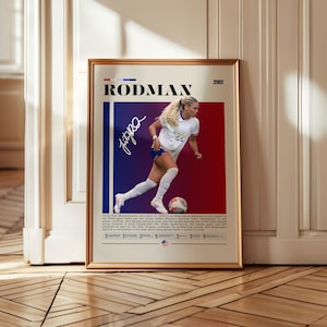 May include: Framed poster of soccer player Trinity Rodman in action. The poster has a red and blue background with the name "RODMAN" at the top. Rodman is wearing a white jersey and blue shorts, with a soccer ball at her feet. The poster includes her signature.