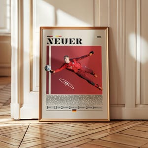 May include: Framed poster featuring a dynamic image of a soccer goalkeeper in a red uniform, mid-air, reaching for a soccer ball. The poster has the name "NEUER" in bold letters, a red background, and text at the bottom.