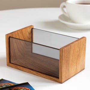 Peut inclure: Une boîte de rangement rectangulaire dotée de deux panneaux latéraux en bois brun clair massif, reliés par une façade et une base en acrylique transparent teinté foncé. Ce contenant ouvert présente une finition en bois naturel et lisse.
