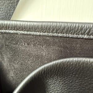 以下が含まれることがあります： 濃いグレーの革製バッグのクローズアップ。 画像は、テクスチャ加工された表面とステッチの詳細を強調しています。 「MADE IN ITALY」の文字が内側の裏地に見えます。