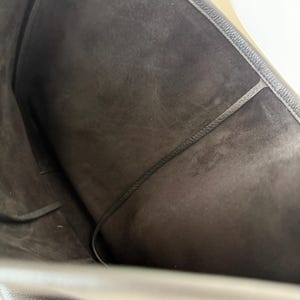 以下が含まれることがあります： 黒いレザートートバッグの内側の様子。内側は濃いグレーのスエードのような素材です。黒いコードが中央を走っています。バッグの縁はコントラストのある糸でステッチされています。バッグは開いており、広々とした内部が明らかになっています。