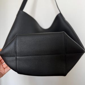 以下が含まれることがあります： 黒い革のショルダーバッグ。幅広のオープントップとシングルショルダーストラップが付いています。バッグは質感のある表面とシンプルでミニマリストなデザインで、日常使いに適しています。